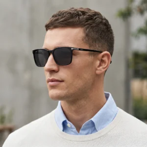 Close-up of a man wearing black sunglasses and a light blue shirt under a cream sweater outdoors.