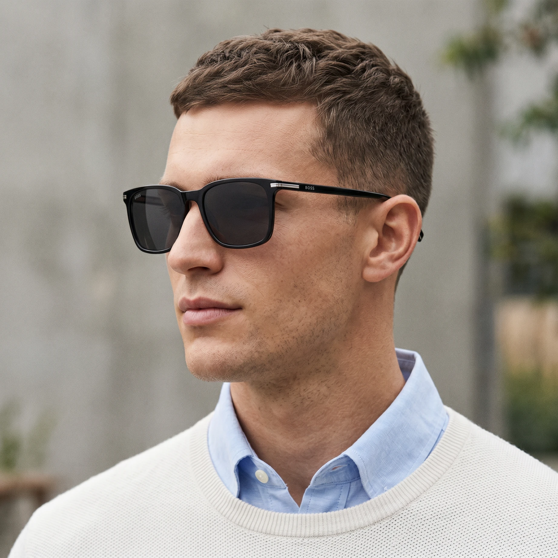 Close-up of a man wearing black sunglasses and a light blue shirt under a cream sweater outdoors.