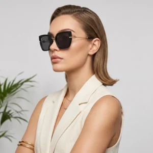 Side profile of a woman wearing oversized black sunglasses and a beige sleeveless blazer, with gold-accented arms.
