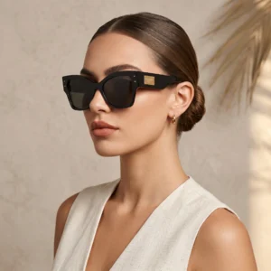 Woman wearing oversized black sunglasses with a gold brand plaque on the temple, neutral background.