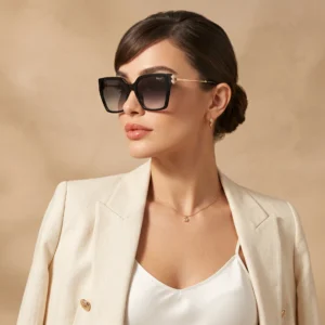 Woman wearing oversized black sunglasses with a beige blazer and pearl earrings, posing against a warm beige backdrop.