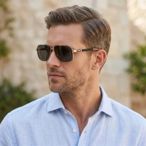 Man wearing brown aviator-style sunglasses and a light blue shirt, outdoors with a blurred stone wall background.