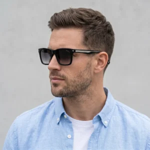 Profile view of a man wearing black sunglasses and a light blue button-down shirt against a gray background, showing part of his face and beard.