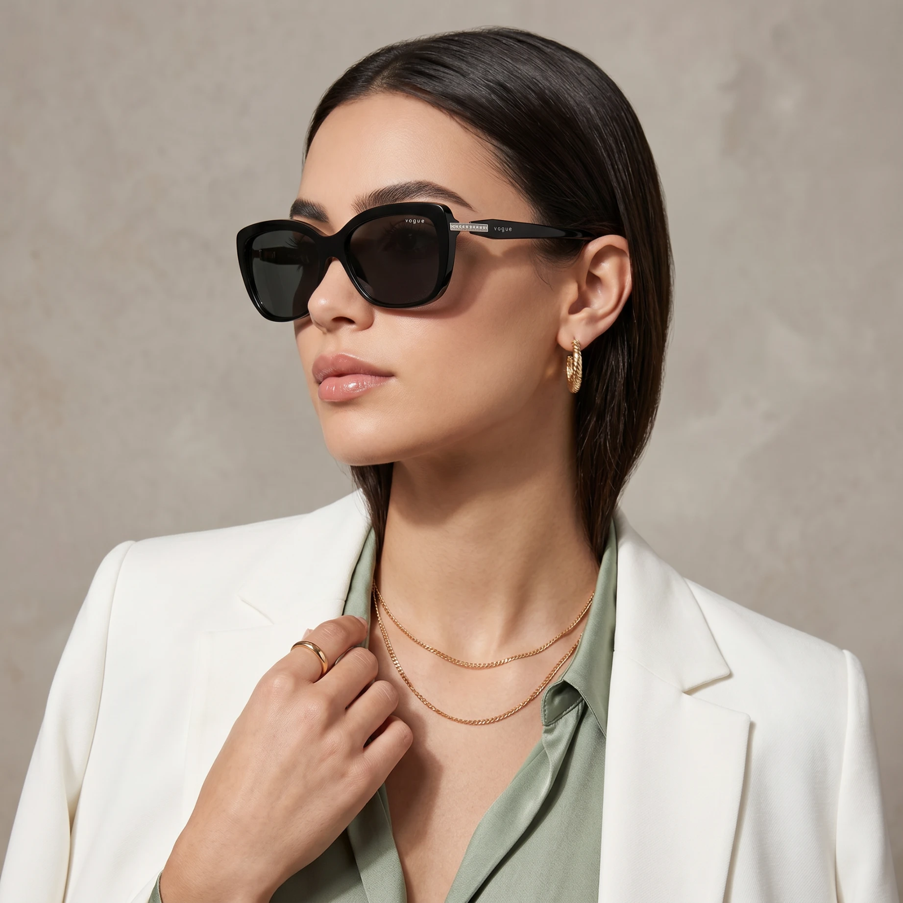 Close-up of a woman wearing oversized black sunglasses, a white blazer, and layered gold necklaces against a neutral backdrop.